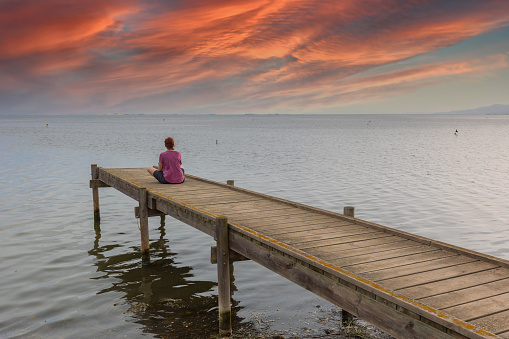 Red-haired woman sitting on a wooden dock watching the sunset.