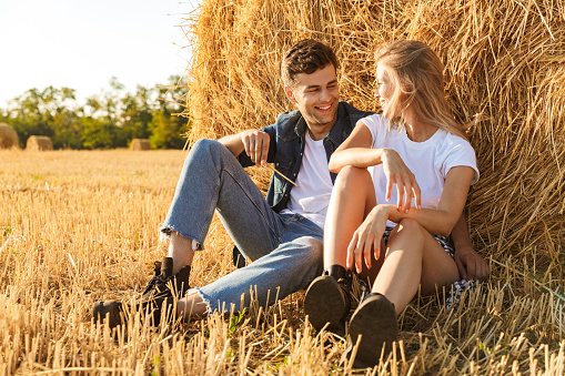 Photo of young couple man and woman sitting under big haystack in golden field during sunny day
