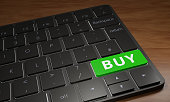 Close up of a computer keyboard on a wooden table. One key is green with the word BUY on it.