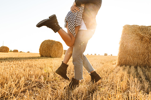 Cropped photo of guy and girl in love walking through golden field with bunch of haystacks and hugging during sunny day