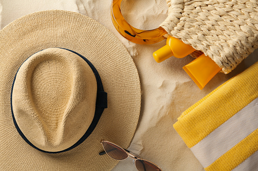 Straw hat, sunglasses, towel and bag with sunscreen on sand, flat lay