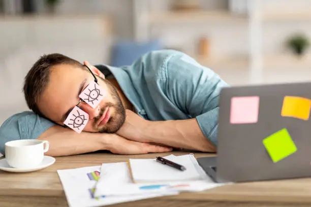 Photo of Lazy unproductive young guy wearing funny sticky notes with open eyes on his glasses, sleeping at workplace