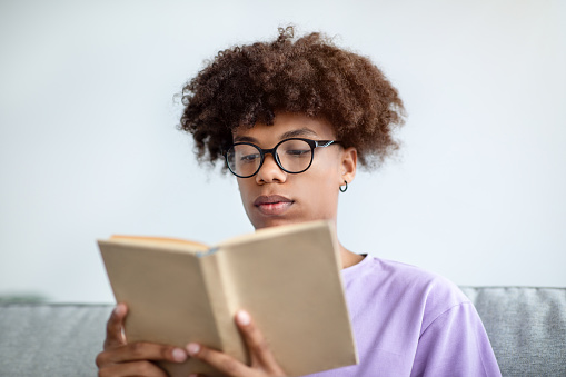 Focused African American teenager reading book, enjoying captivating story at home. Intelligent black teen student with textbook getting ready for test or exam, studying remotely indoors
