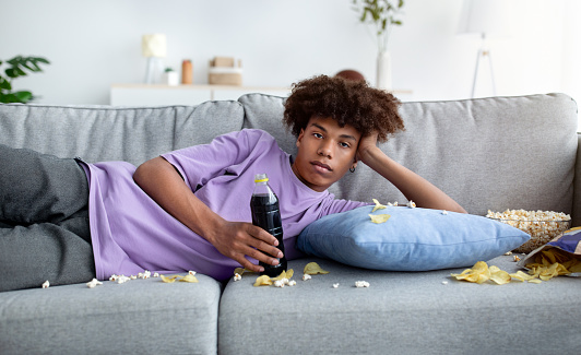 Bored black teenager lying on couch with scattered food, watching dull show or movie on TV, killing time at home. African American adolescent being lazy, eating snacks, staring into television