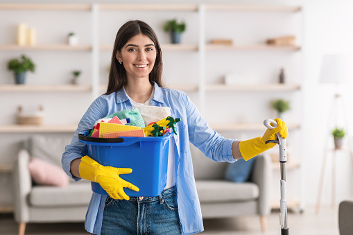 Portrait of smiling millennial lady holding bucket with cleaning supplies and mop, posing and looking at camera standing in living room. Professional cleaning service specialist wearing rubber gloves