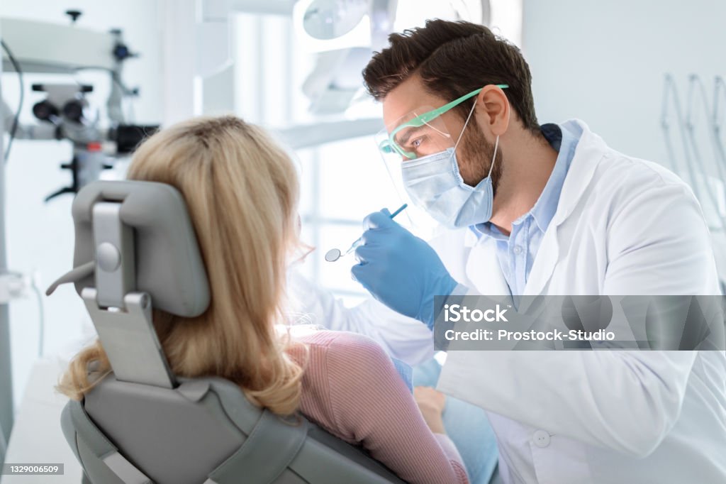 Man dentist in face mask and glasses doing treatment Man dentist in face mask and glasses doing treatment for patient blonde lady, holding dental tools, wearing rubber gloves. Stomatology, dentistry, modern dental clinic concept Dentist Stock Photo