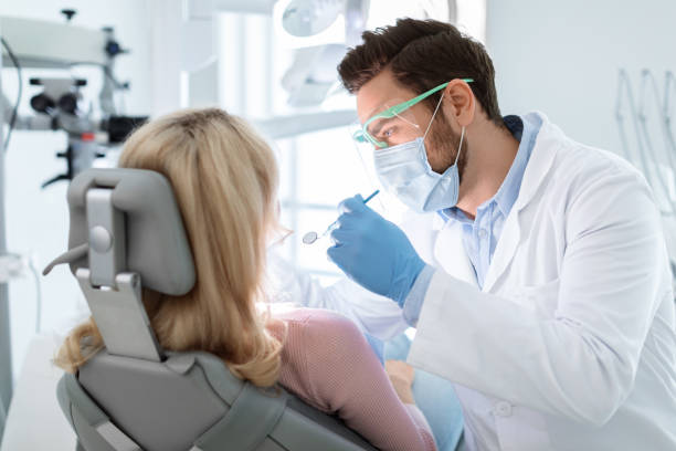 Man dentist in face mask and glasses doing treatment Man dentist in face mask and glasses doing treatment for patient blonde lady, holding dental tools, wearing rubber gloves. Stomatology, dentistry, modern dental clinic concept dentist photos stock pictures, royalty-free photos & images