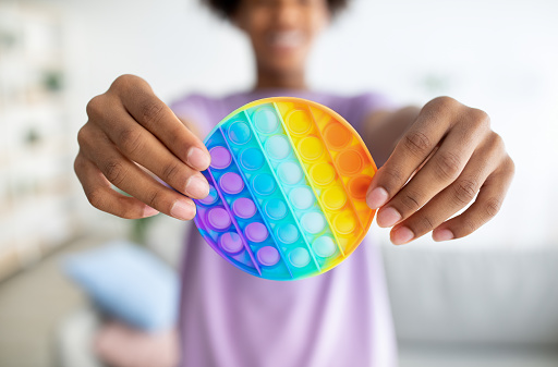Cropped view of black teenager holding POP IT toy in outstretched hands at home, closeup. Unrecognizable African American youth playing with modern sensory antistress trinket