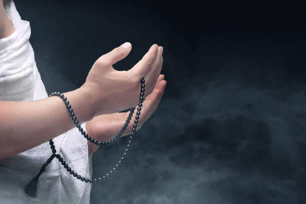 A Muslim man in ihram clothes praying with prayer beads on his hands with colored background