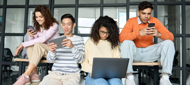 Multiracial students at high school campus using devices. Course preparation. Young diverse multiracial cool group of high school college students girls and guys sitting at university campus space holding technology gadgets using digital devices studying together. letter z stock pictures, royalty-free photos & images