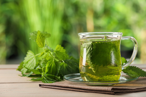 Glass cup of aromatic nettle tea and green leaves on wooden table outdoors, space for text