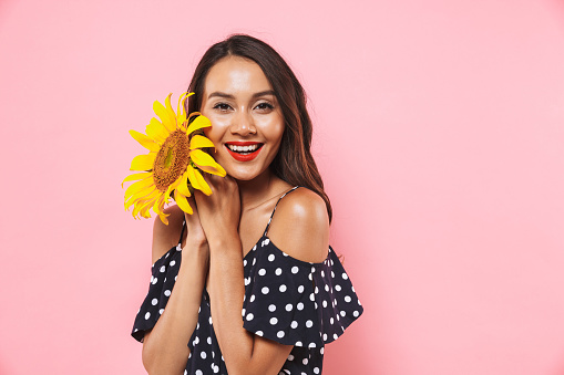 Happy brunette woman in dress posing with sunflower and looking at the camera over pink background