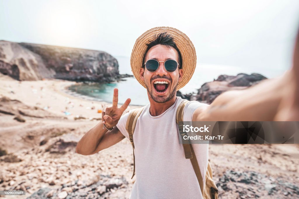 Junger Mann mit Rucksack macht Selfie-Porträt auf einem Berg - Lächelnder glücklicher Kerl, der Sommerferien am Strand genießt - Millennial zeigt Siegeshände Symbol in die Kamera - Jugend und Reise - Lizenzfrei Reise Stock-Foto