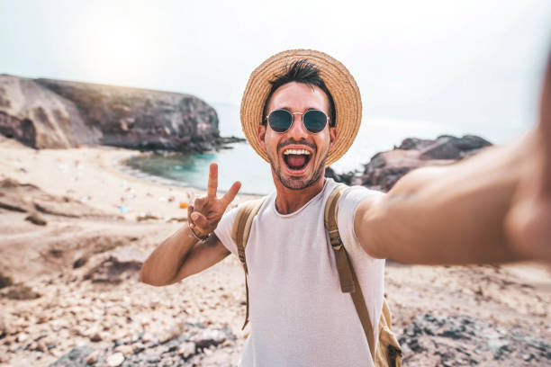 Young man with backpack taking selfie portrait on a mountain - Smiling happy guy enjoying summer holidays at the beach - Millennial showing victory hands symbol to the camera - Youth and journey stock photo