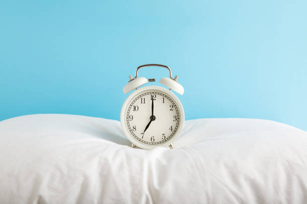 white alarm clock on pillow at light blue wall background. pastel color. 7 o'clock in morning. waking time concept. closeup. front view. - pillow imagens e fotografias de stock
