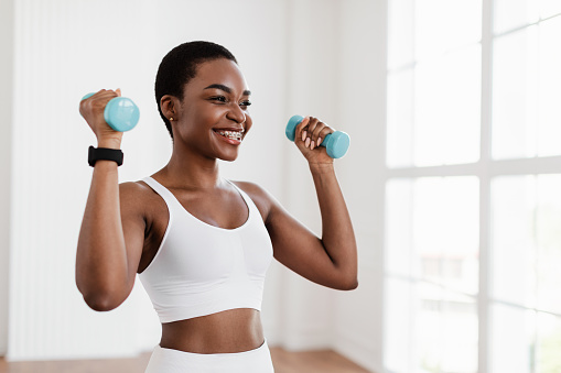 Fitness And Sport Concept. Portrait Of Smiling Young Black Woman In Bra Exercising With Two Dumbbells Posing At Gym, Enjoying Workout. Selective Focus, Blurred Background, Free Copy Space