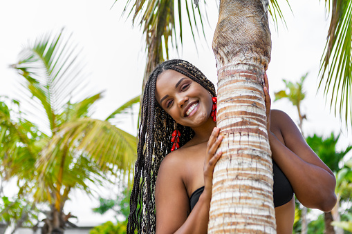 African American woman in summer clothes behind a palm tree on the beach. Cheerful woman on the beach smiling and looking at camera.