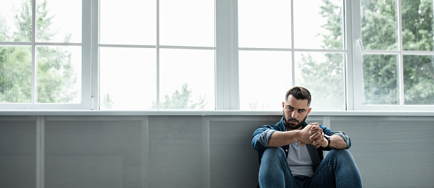 Man feeling sad, tired and worried, suffering depression in mental health, problems and broken heart. Frustrated young handsome bearded guy sitting on floor near large window, copy space, panorama