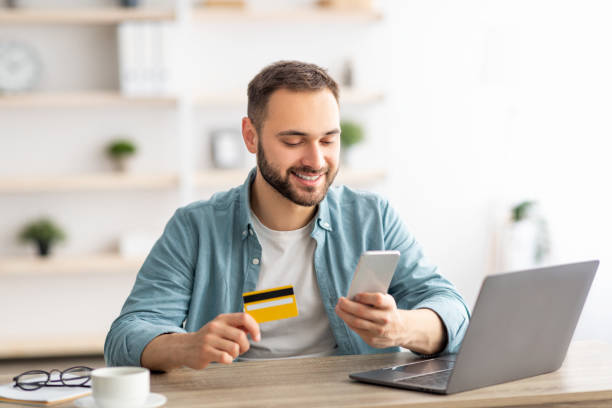 Happy Caucasian man buying things online, using smartphone, laptop and credit card, enjoying shopping in internet Happy Caucasian man buying things online, using smartphone, laptop and credit card, enjoying shopping in internet. Positive young guy purchasing goods on web, making remote payment person paying stock pictures, royalty-free photos & images