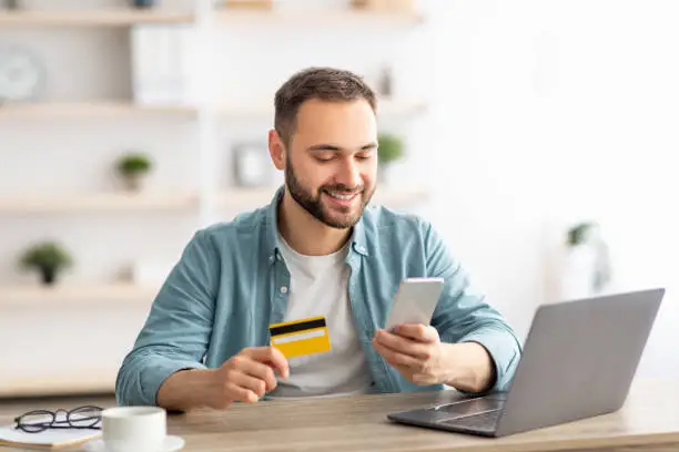 Photo of Happy Caucasian man buying things online, using smartphone, laptop and credit card, enjoying shopping in internet