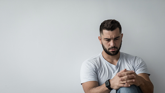 Negative human emotions, deep depression, loneliness, sadness, stress and psychological problems. Serious young handsome muscular bearded guy suffer from anxiety, on gray wall background, copy space