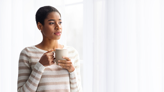 Young Black Lady Drinking Coffee While Standing Near Window At Home, Relaxed Millennial African American Woman Holding Cup In Hands, Enjoying Hot Drink And City View, Panorama With Copy Space