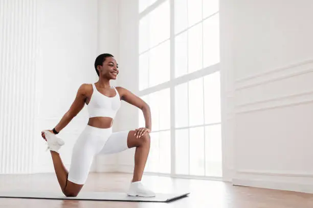 Warming Up Before Workout. Smiling Sporty Black Woman Training At Home Or Fitness Club Studio, Beautiful Female Stretching Legs After Exercises On Mat, Wearing White Bra And Bike Shorts, Free Space