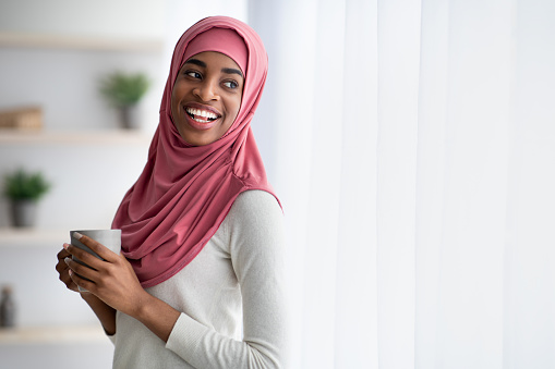 Happy Smiling Black Muslim Woman In Hijab Drinking Coffee Near Window At Home, Positive African Islamic Female Holding Cup With Hot Drink And Looking Away, Enjoying Domestic Leisure, Copy Space