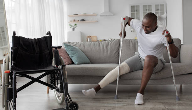 Patient is recuperating in living room due to illness Patient is recuperating in living room due to illness. Concentrated cheerful tense adult african american guy with broken leg in cast stands up with crutches from couch near wheelchair, panorama orthopedic cast stock pictures, royalty-free photos & images