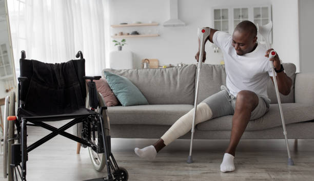 Patient is recuperating in living room due to illness Patient is recuperating in living room due to illness. Concentrated cheerful tense adult african american guy with broken leg in cast stands up with crutches from couch near wheelchair, panorama injured stock pictures, royalty-free photos & images