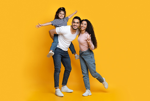 Cheerful Middle Eastern Family Of Three Having Fun Together Over Yellow Background, Happy Young Arab Parents And Their Little Daughter Laughing And Smiling At Camera, Full Length Shot, Copy Space