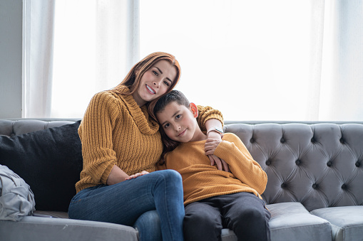 Average age 30-year-old Latin mother is sitting on the sofa at home while being hugged from behind by her son is being hugged by her 12-year-old Latino teenage son both dressed alike in yellow jackets