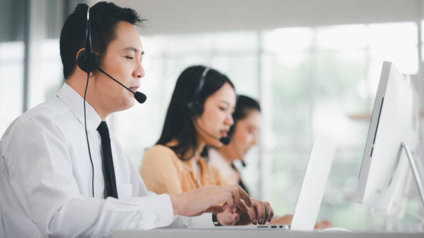 equipe de call center na ásia usando fones de ouvido com microfones. estresse e dores de cabeça enquanto atendem clientes em mesas e computadores. conceito de serviço e consultoria. conceito de comunicação. - neutral look audio - fotografias e filmes do acervo