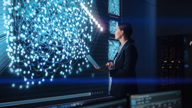 istock Young Female Computer Science Engineer Working on Laptop next at Big Screen Display Showing Neural Data. Telecommunications Technology Company System Control and Monitoring Room with Servers. 1329049647