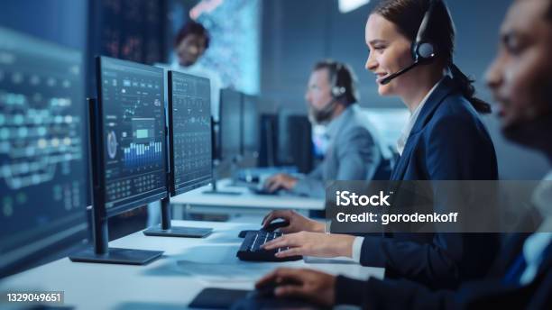 Happy Beautiful Technical Customer Support Specialist Having A Headset Call While Working On A Computer In A Dark Monitoring And Control Room Filled With Colleagues And Display Screens Stock Photo - Download Image Now