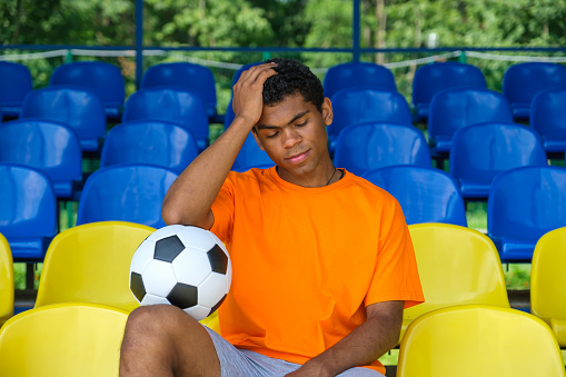 Young Brazilian guy with a soccer ball sitting on an empty football grandstand outdoor in summer