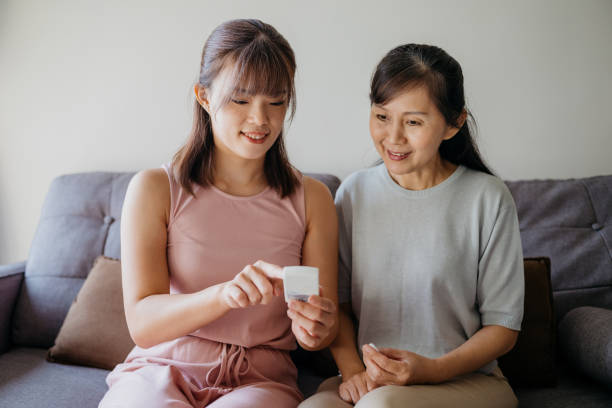 Asian woman helping her mother check blood sugar level using a blood glucose meter at home Image of an Asian Chinese woman helping her mother check blood sugar level using a blood glucose meter at home. Young woman showing her mother the test result on the device. blood sugar stock pictures, royalty-free photos & images