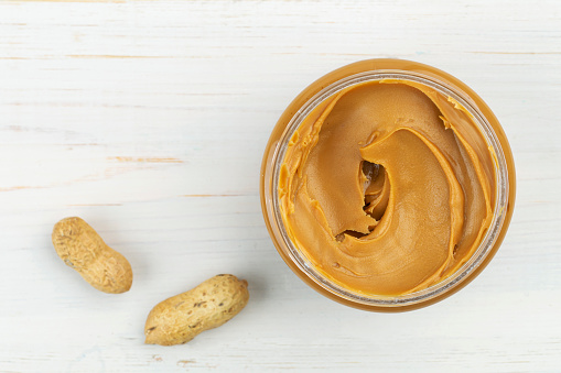 Jar of peanut butter on a white wooden table with peanuts scattered nearby. Space for text. Top view.