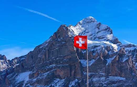 The Swiss flag flutters and tourists admire the Monch and Jungfrau peaks in Grindelwald, Switzerland.