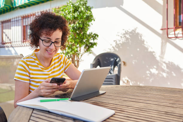 Young business woman telecommuting at an outdoor site Caucasian curly-haired woman teleworking using her mobile phone and digital tablet outdoors digital nomad stock pictures, royalty-free photos & images