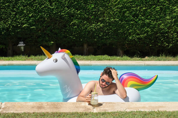 istock Young man wearing sunglasses combing with his fingers drinking lemonade into a unicorn inflatable ring in a swimming pool. Summer concept. 1329081449