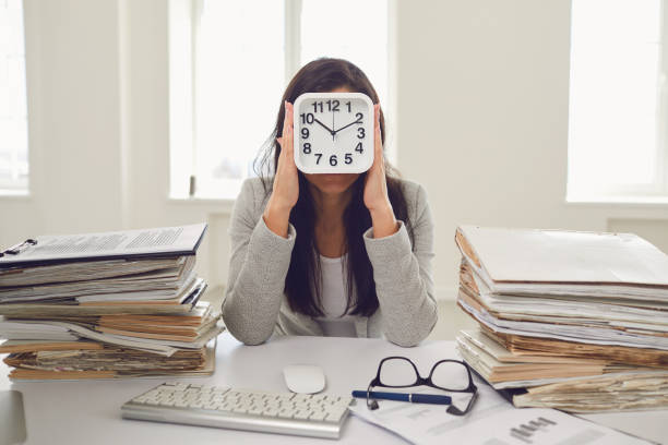 femme d’affaires tenant un réveil devant son visage tout en étant assise à une table dans le bureau - clock face clock time deadline photos et images de collection