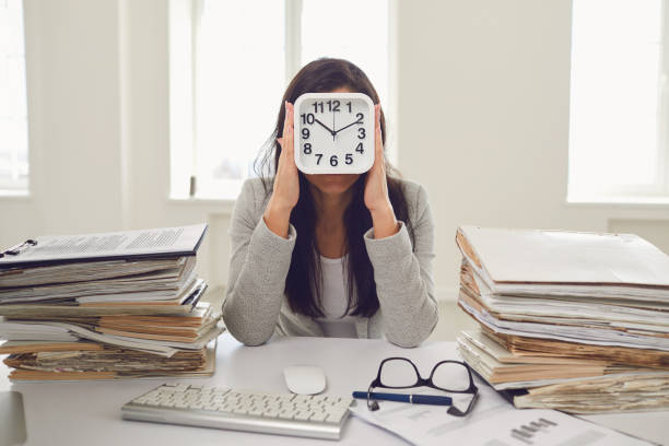 business woman holding an alarm clock in front of her face while sitting at a table in the office - clock face fotos imagens e fotografias de stock