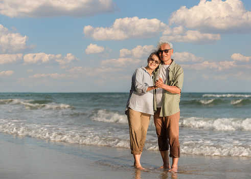 Asian senior couple tourist is hand in hands, embrace and dancing on tropical sea beach in summer vacation. Happy family couple elderly older retire resting relax together lifestyle on coast outdoor.