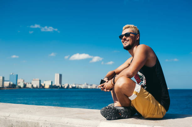 istock Tourist man sitting on stone while smiling and posing against blue sea 1329107485