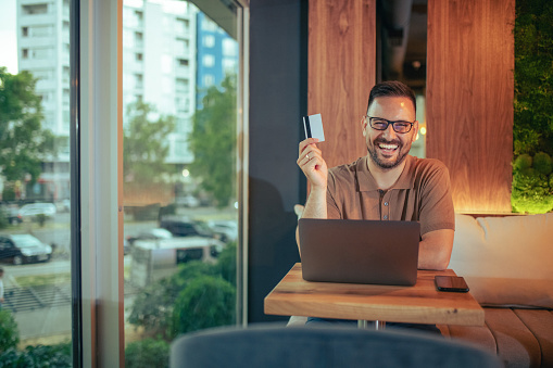 An image portraying a handsome young businessman enjoying the after hours in his favorite cafe. He's posing with a credit card he just made an online purchase with.