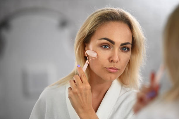 istock Gorgeous young woman looking at her mirror reflection and massaging her face with jade roller stone massager, performing lymphatic drainage facial massage 1329108649