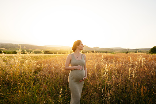 Photo of a pregnant woman standing alone in a field, stroking her baby bump and enjoying her time in nature.