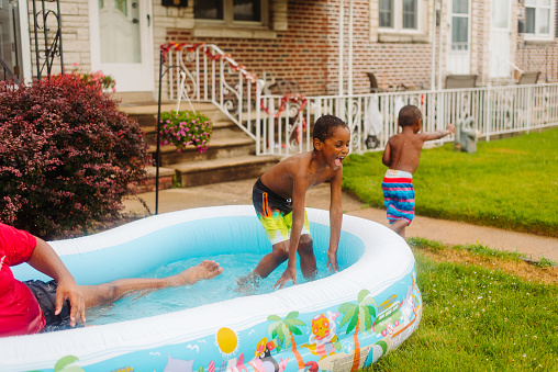 Black/African-American boys ages toddler and child are playing in the pool with their father during the summer. Colorful and filled with splashing, laughter, and joy.