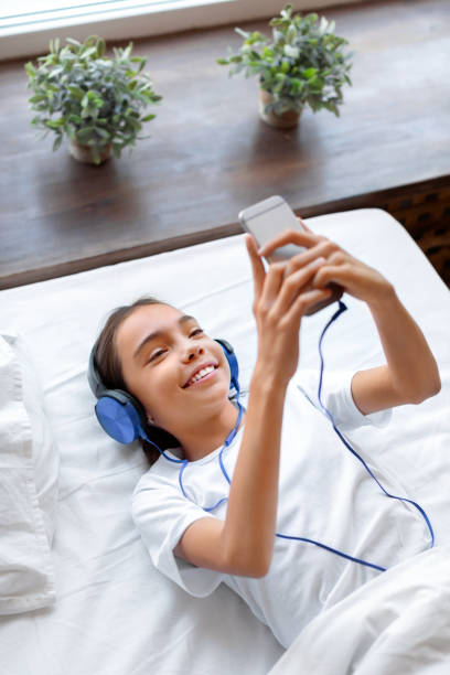 pionowe zdjęcie nastoletniej dziewczyny w łóżku ze smartfonem podczas rozmowy wideo online. komunikuje się z przyjaciółmi lub słucha muzyki przez słuchawki - video game friendship teenager togetherness zdjęcia i obrazy z banku zdjęć